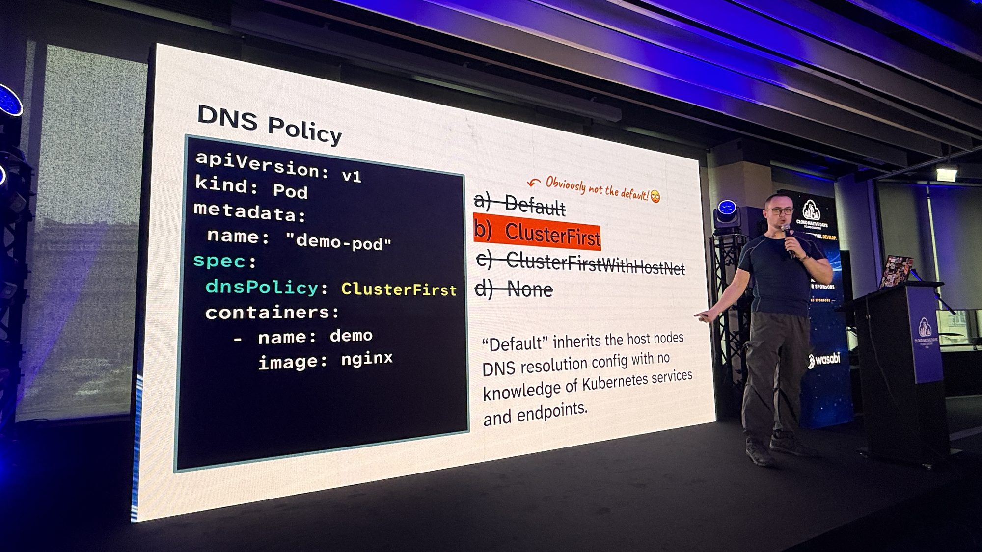 Marcus Noble pointing at a slide titled DNS Policy, with ClusterFirst highlighted and the note: Obviously not the default