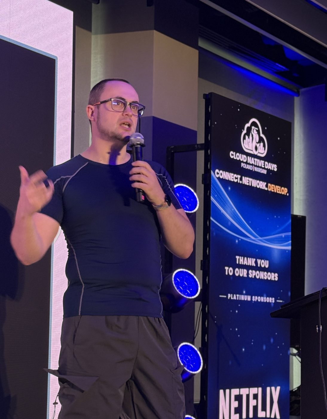 Marcus Noble on stage with a microphone in front of a Cloud Native Days Poland sponsor banner