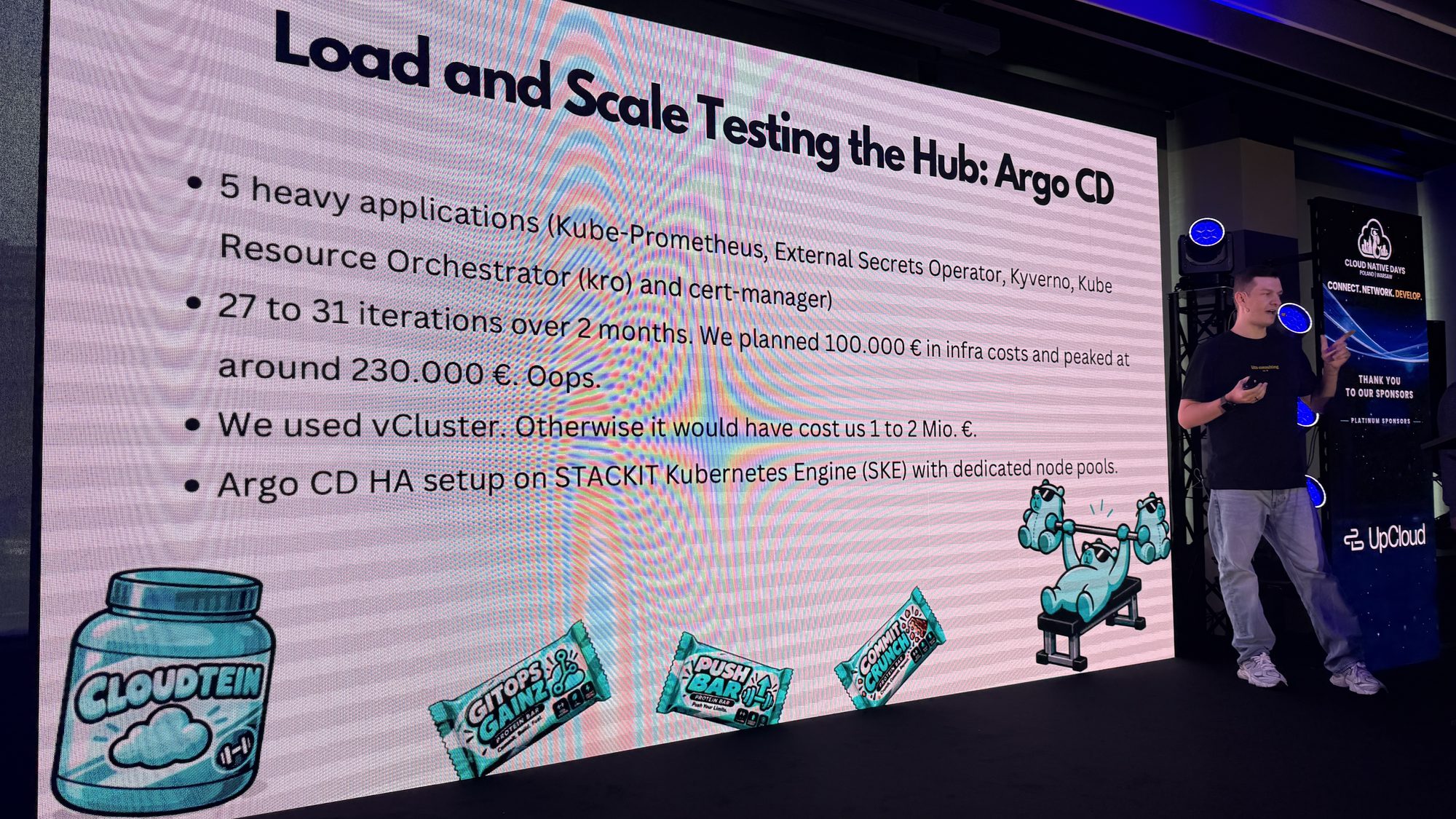 Artem Lajko next to a slide titled Load and Scale Testing the Hub: Argo CD, decorated with Cloudtein protein powder, GitOps Gainz protein bars and a weightlifting bear
