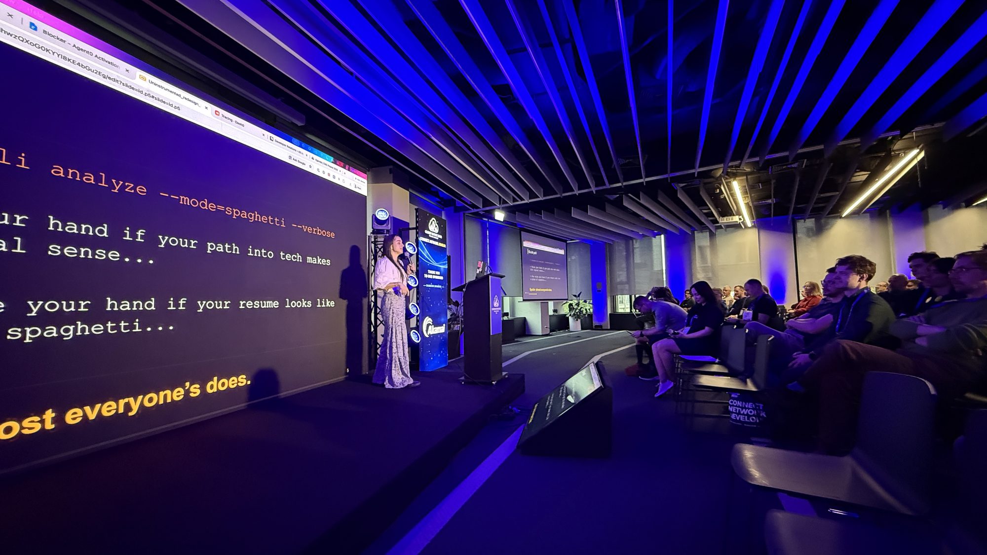 Julia on stage with the audience in view, next to a slide asking people to raise their hand if their path into tech makes linear sense or if their resume looks like spaghetti