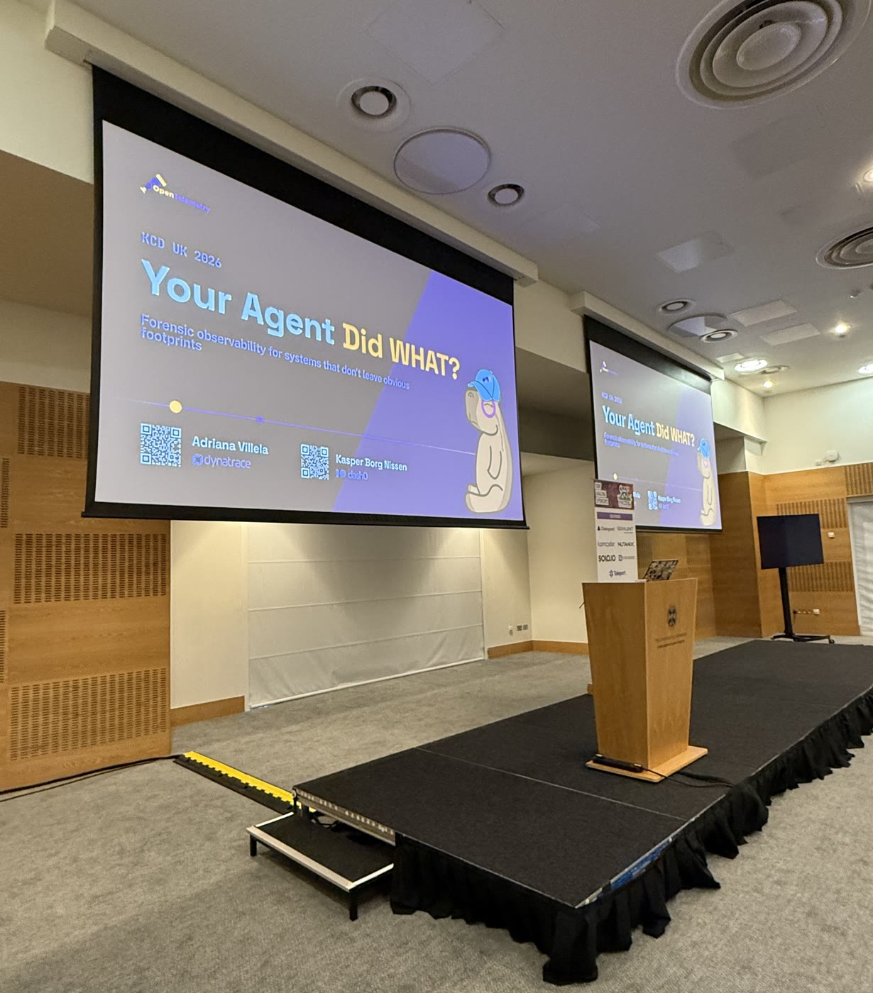 An empty stage with the Your Agent Did WHAT? title slide for KCD UK 2026 on both screens