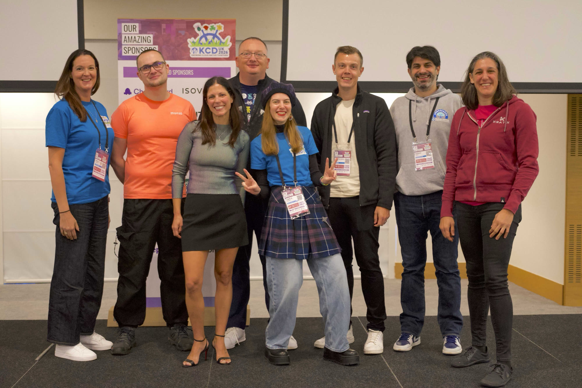 Eight CNCF Ambassadors, including Adriana, Koray, Abby and Kasper, in front of the KCD UK 2026 sponsor banner
