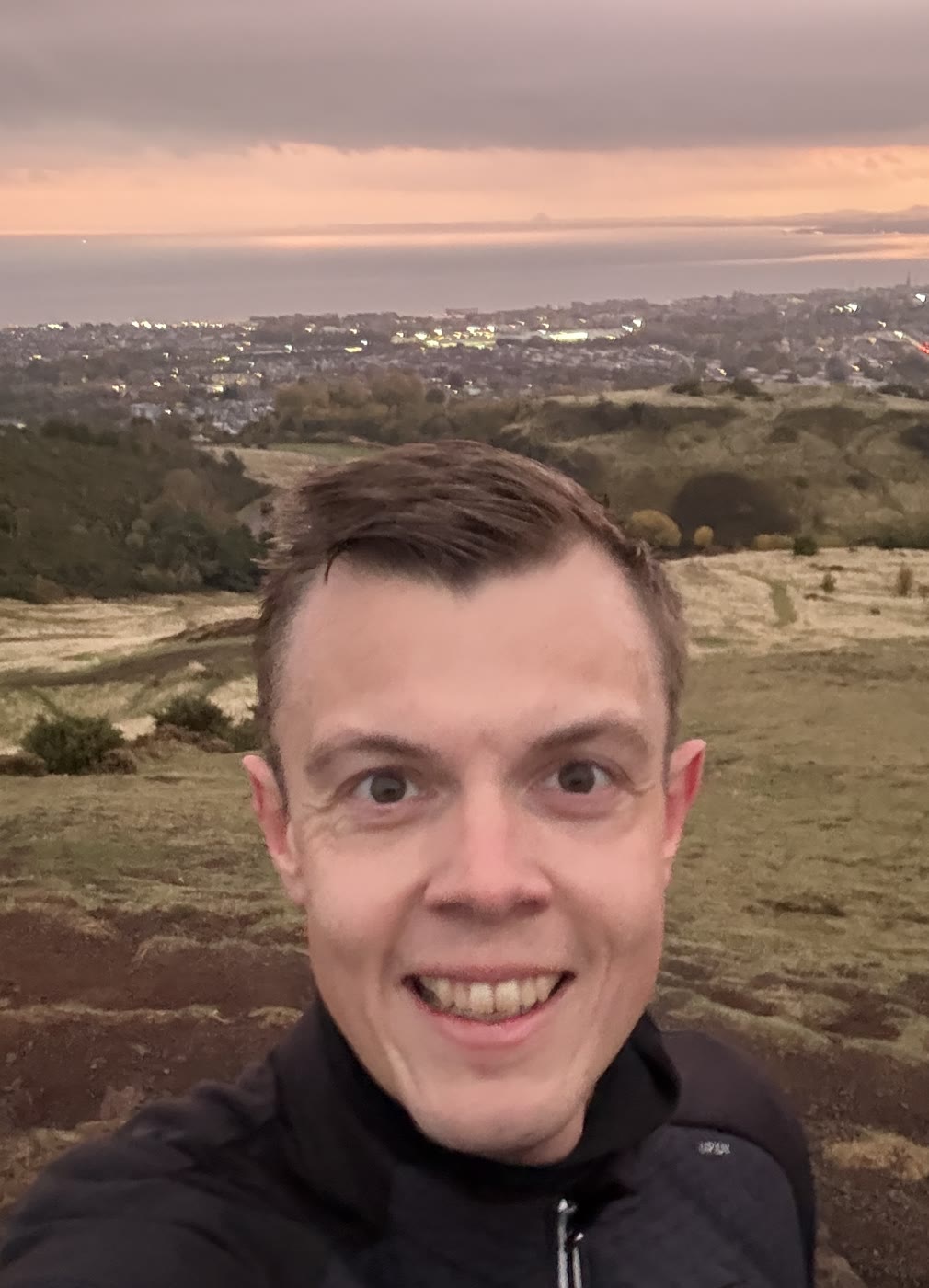 Kasper in a black running jacket taking a selfie on Arthur's Seat, with Edinburgh and the sea behind at dusk