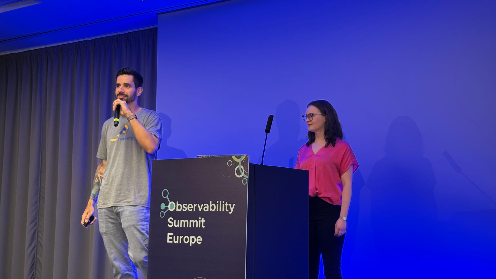 Juliano Costa in an OpenTelemetry t-shirt speaking into a microphone next to Johanna Öjeling