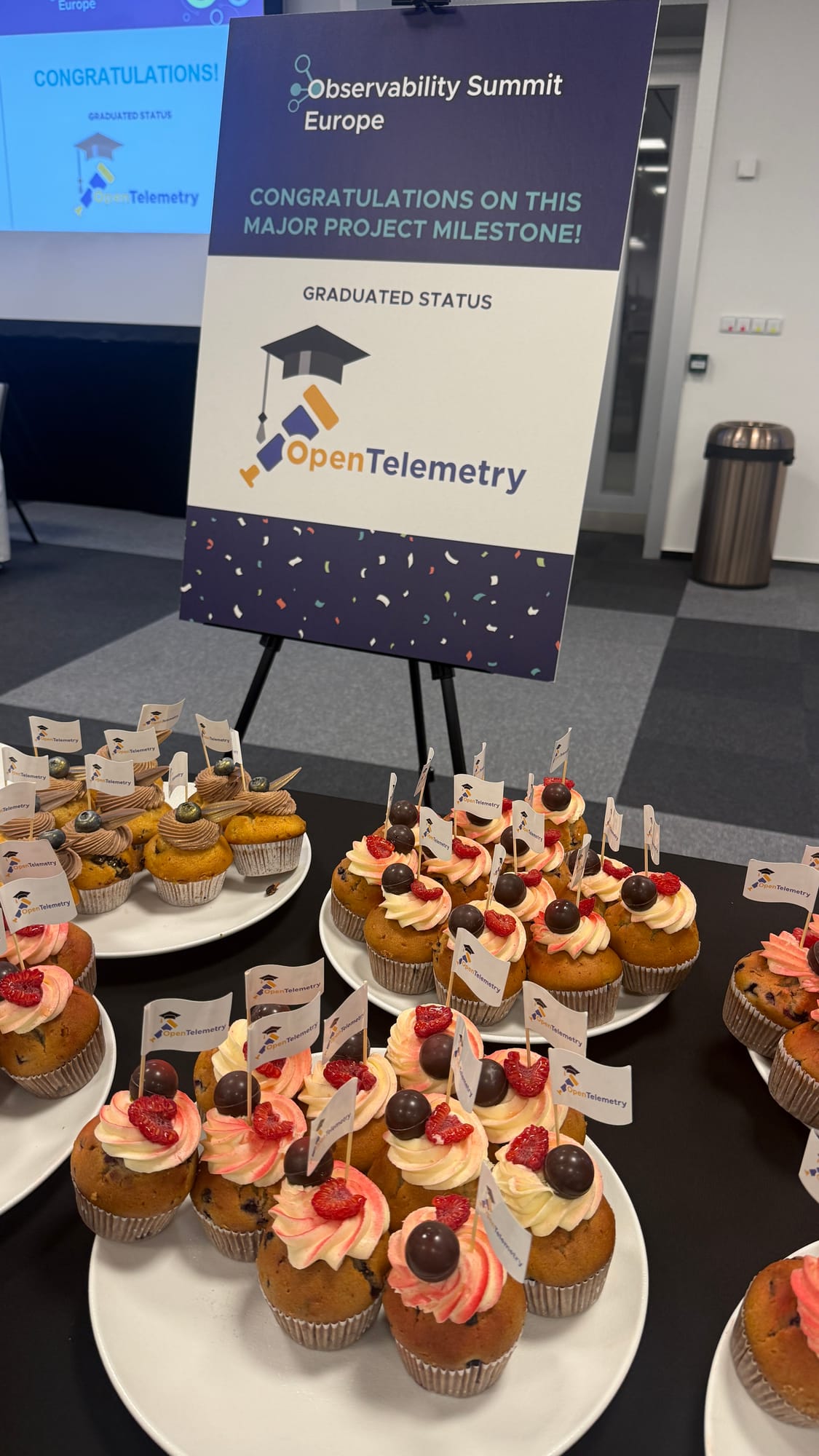 Cupcakes with OpenTelemetry flags in front of a sign congratulating OpenTelemetry on graduated status