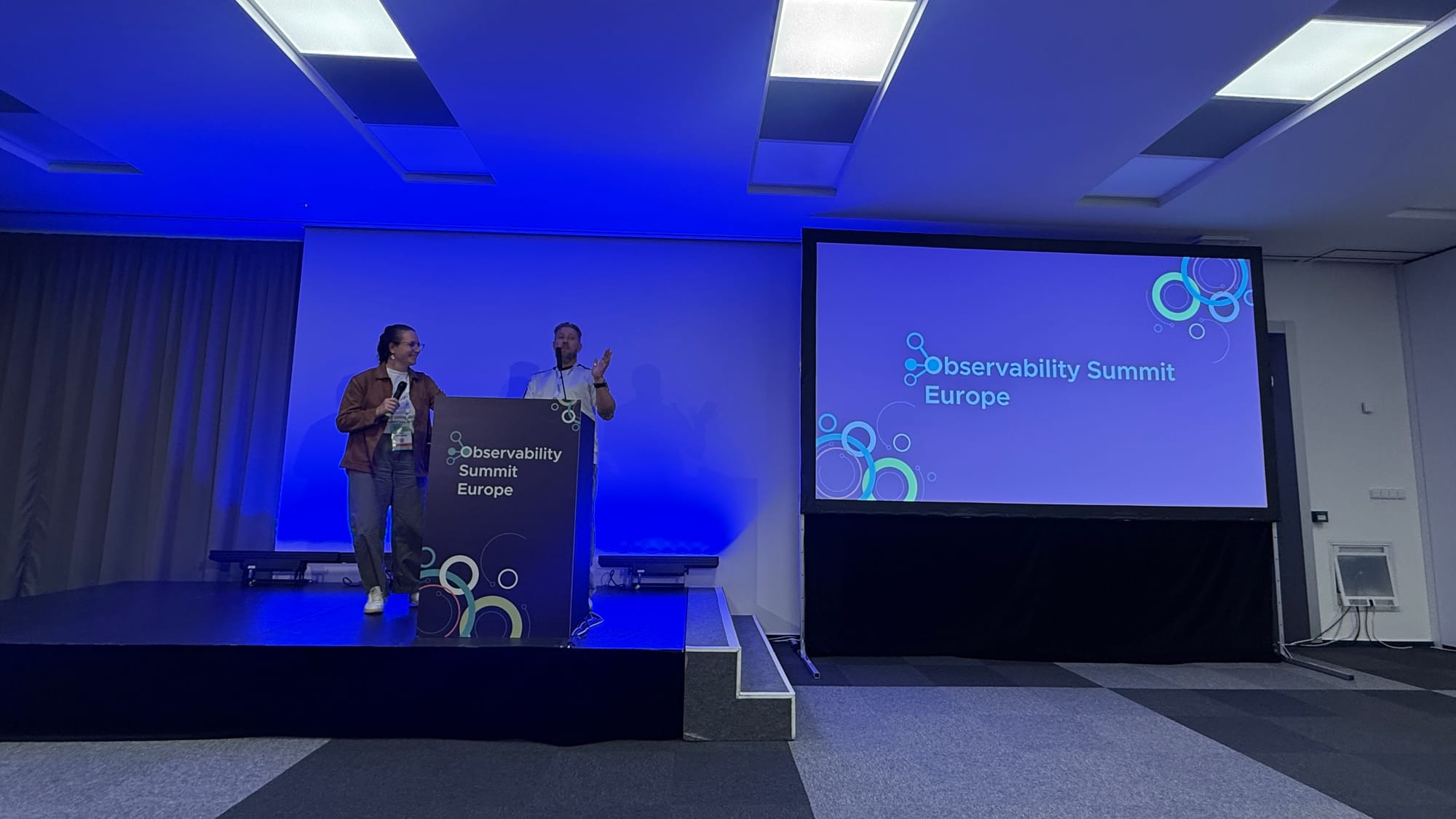 Iris Dyrmishi and Henrik Rexed on stage next to the Observability Summit Europe title slide
