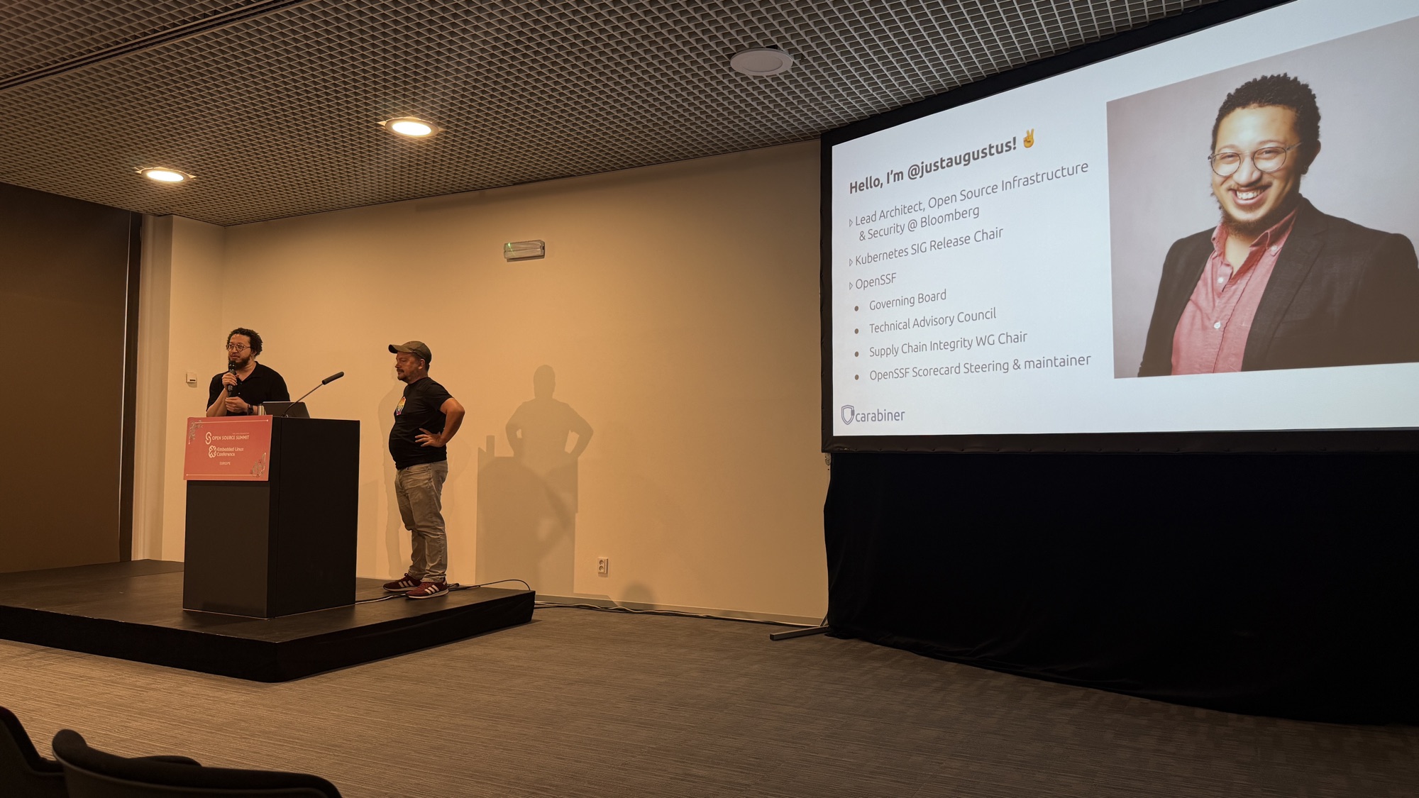 Stephen Augustus and Adolfo García Veytia on stage next to Stephen's intro slide listing Bloomberg, Kubernetes SIG Release and OpenSSF roles